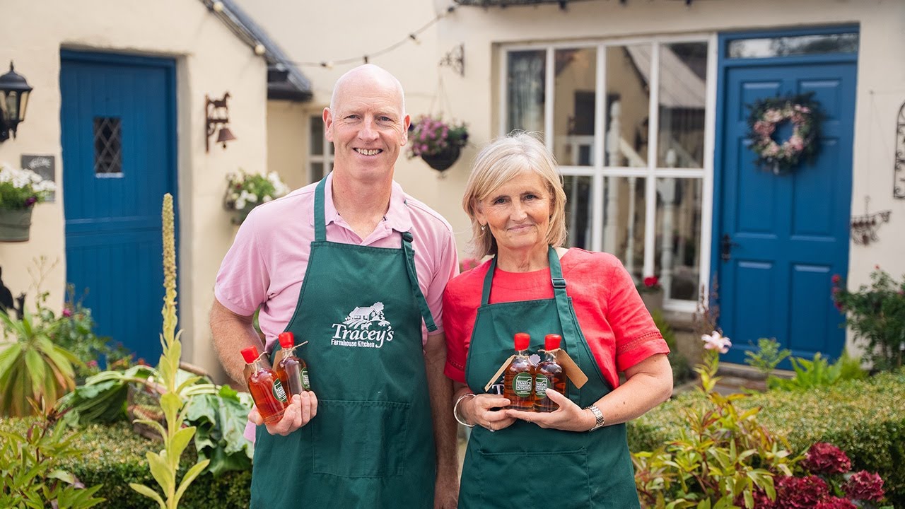 Two people in aprons holding bottles in front of a house with blue doors.